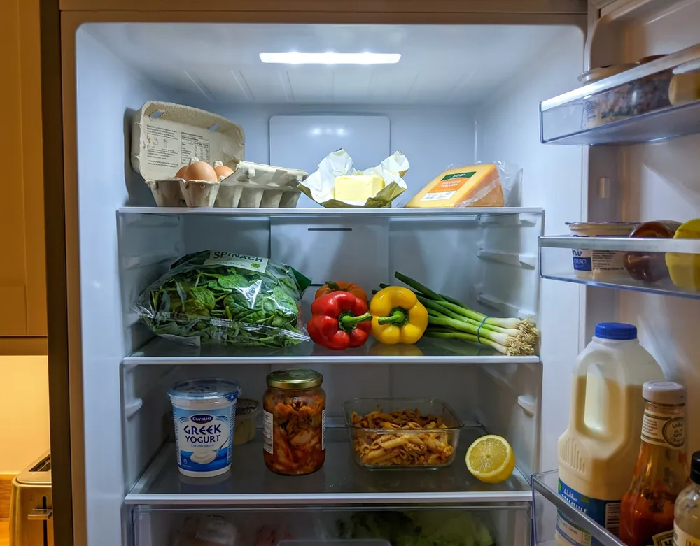 The inside of an open home fridge holding eggs, butter, cheddar, spinach, peppers, spring onions, yoghurt and leftovers